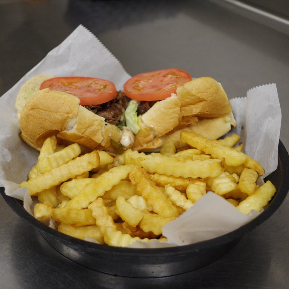 Two cheesesteak sandwiches with tomatoes and lettuce, served with crinkle-cut fries in a black basket.